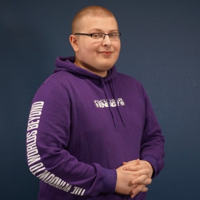 Henkilökuva, luhyet hiukset, ohutsankaiset silmälasit, violetti huppari, kädet yhdessä edessä, tummansininen tausta.